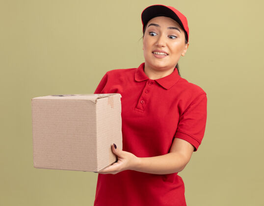 制服年轻的送货员身穿红色制服 戴着帽子 手里拿着一个纸板箱 站在绿色的墙上 又高兴又惊讶地看着它送货站着帽子