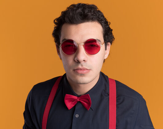 男人带着蝴蝶结戴着眼镜和吊带的时髦男人站在橙色的墙上 带着怀疑的表情看着前面时尚领带穿着