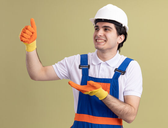 建筑工人年轻的建筑工人身穿建筑制服 戴着橡胶手套戴着安全帽 站在绿色的墙壁上 一边看一边竖起大拇指 愉快地微笑着制服橡胶手套