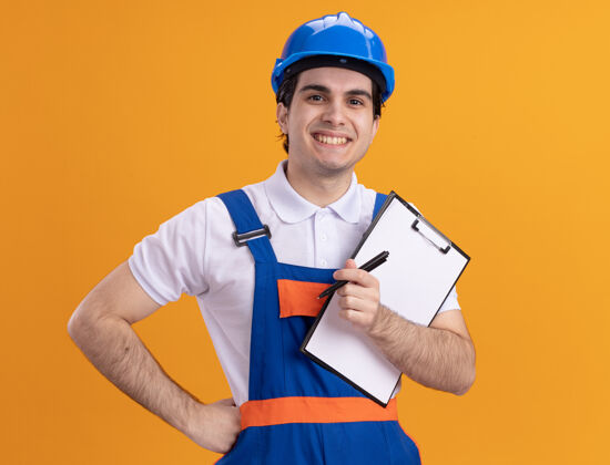 头盔年轻的建筑工人身穿建筑制服 戴着安全帽 拿着写字板和钢笔 站在橘色的墙上 面带微笑地看着前方钢笔欢呼制服