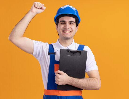 男人年轻的建筑工人穿着建筑制服 戴着安全帽 拿着写字板看着前面 高兴而积极地举起拳头站在橙色的墙上提高立场建设者