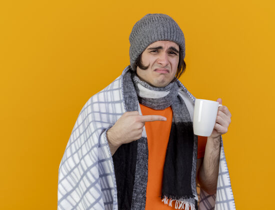 不愉快一个戴着冬天帽子 围巾裹着格子布的年轻病患 指着一杯孤立在橙色背景上的茶年轻拿着格子