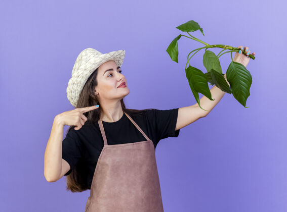 园艺穿着制服的漂亮园丁女孩戴着园艺帽 抬起头来指着隔离在蓝色背景上的植物女孩帽子制服