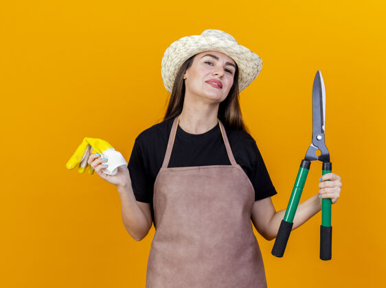 微笑微笑美丽的园丁女孩穿着制服 戴着园艺帽拿着剪刀 手套隔离在橙色背景上花园帽子手握