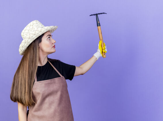 提高美丽的园丁女孩穿着制服 戴着园艺帽和手套 抬起头 看着在蓝色背景上与复制空间隔离的耙子园艺耙子手套