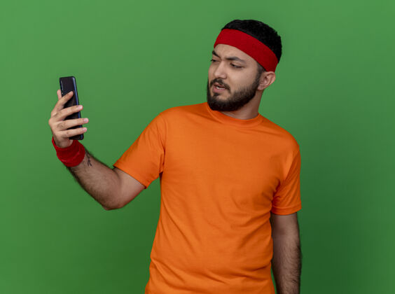 年轻戴着头带和腕带 拿着手机 看着绿色背景上孤立的手机 一个未被释放的运动型年轻人运动头带不愉快