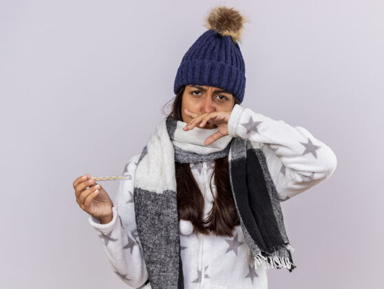 手悲伤的生病的年轻女孩戴着冬天的帽子 戴着围巾 拿着温度计 用手擦鼻子 背景是白色的女孩围巾体温计