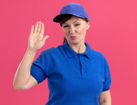 年龄身穿蓝色制服 头戴鸭舌帽的中年女送货员自信地看着前面 手张开站在粉红色的墙上女人立场帽子