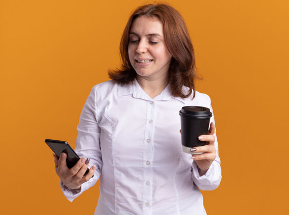 衬衫身穿白衬衫 手持智能手机 手持咖啡杯 面带微笑地看着手机的年轻女子站在橙色的墙上脸站着拿着