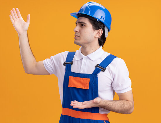 建筑工人年轻的建筑工人穿着建筑制服 戴着安全帽 一边看一边不高兴地用手站在橙色的墙上制服站立建筑