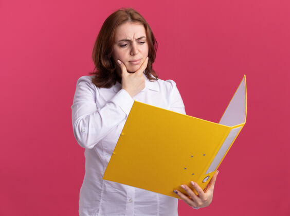 衬衫穿着白衬衫的年轻女子拿着文件夹 困惑地看着它 困惑地站在粉红色的墙上困惑拼图文件夹