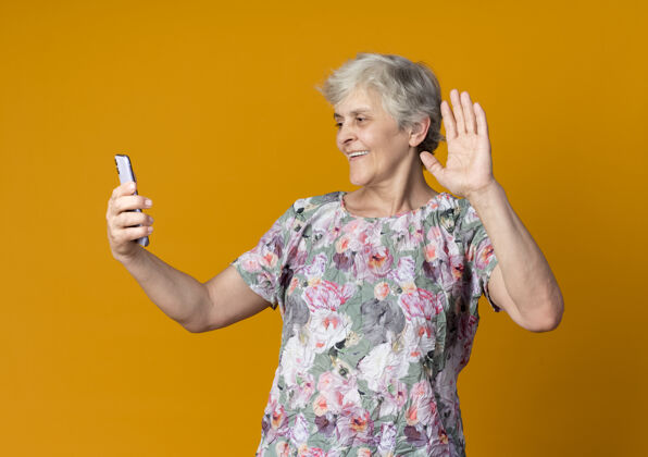 举起来微笑的老妇人举起手 看着隔离在橙色墙上的电话老人微笑抱着