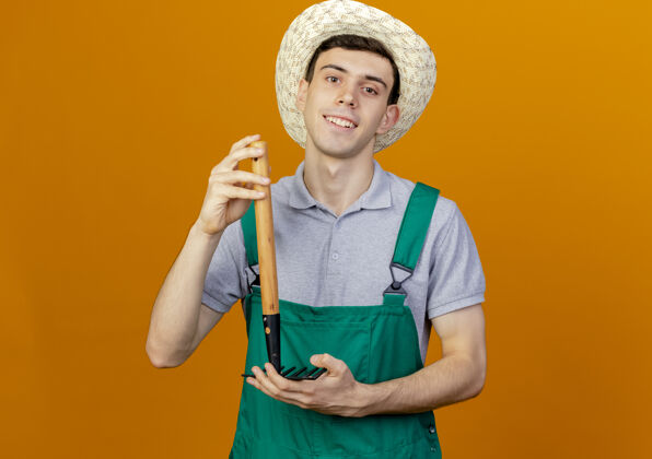 年轻微笑着的年轻男园丁戴着园艺帽 把耙子举得上下颠倒微笑花园帽子