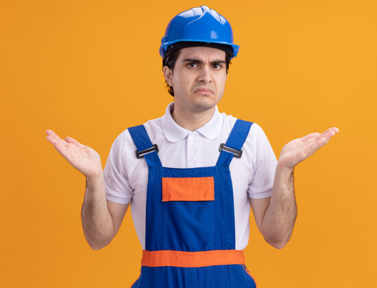 手臂年轻的建筑工人穿着建筑制服 戴着安全帽 站在橙色的墙上 困惑地看着前方 不高兴地举起双臂困惑不高兴站立