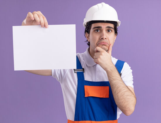 安全年轻的建筑工人穿着建筑制服 戴着安全帽 拿着一张空白的纸 看着前面 愁眉苦脸地撅着嘴唇站在紫色的墙上表情空白钱包
