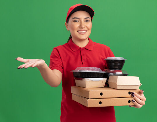 制服身穿红色制服 头戴鸭舌帽的年轻女送货员手持披萨盒和食品包 面带微笑 抬起手臂 站在绿色墙壁上做欢迎手势递送微笑盒子