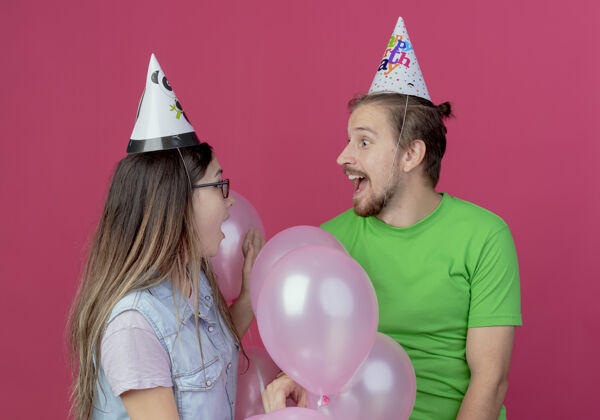 穿一对戴着派对礼帽的年轻夫妇惊讶地看着彼此站在粉红色墙上的氦气球年轻情侣生日