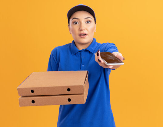 表演身穿蓝色制服 头戴鸭舌帽 拿着披萨盒的年轻女送货员站在橘色的墙上 看着前面的智能手机 既惊讶又困惑送货站着拿着