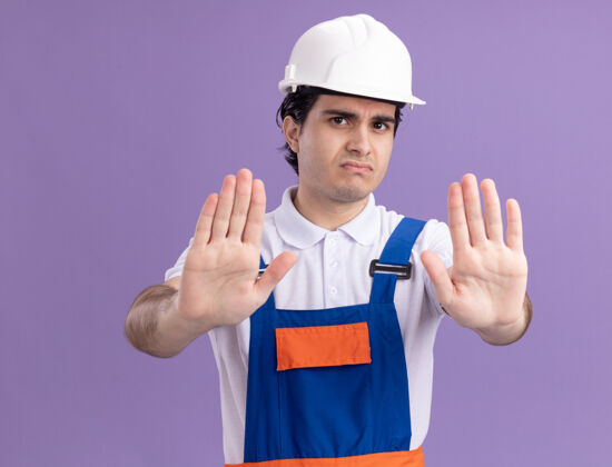 脸年轻的建筑工人身穿建筑制服 头戴安全帽 表情严肃 双手站在紫色的墙上 做着停车的手势年轻严肃停车