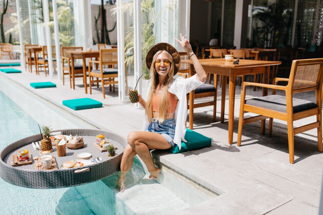乐趣苗条兴奋的金发女人在度假胜地摆姿势夏天美丽的女人在游泳池附近享受水果和鸡尾酒的肖像棕榈树帽子太阳