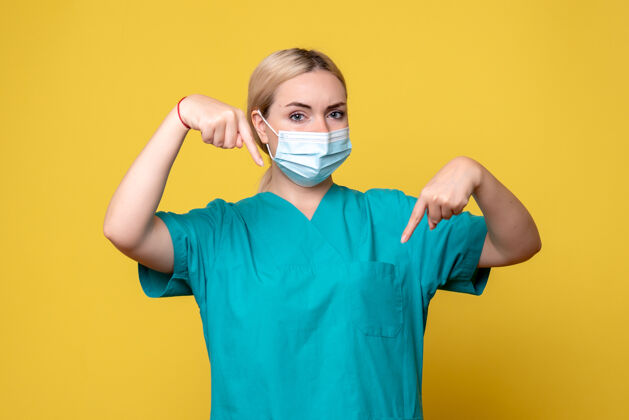 成人黄色墙壁上穿着医用衬衫和无菌口罩的年轻女医生的正面图正面大流行护士
