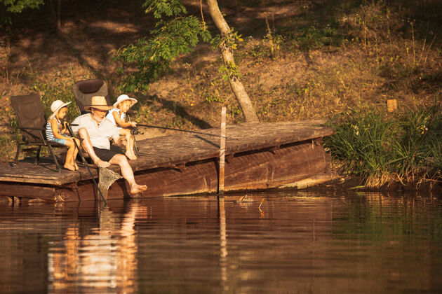 男可爱的小女孩和他们的爷爷在湖边或河边钓鱼在夏天的日落时分 在靠近水和森林的码头上休息家庭 娱乐 童年 自然的概念年龄女孩兴奋