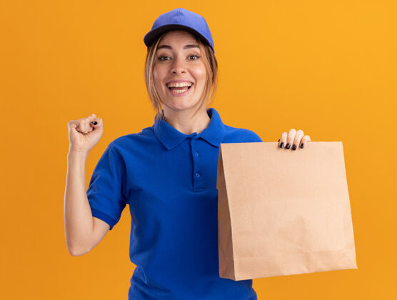 送货快乐的年轻漂亮的女送货员穿着制服 握紧拳头 拿着橙色的纸包年轻姿势漂亮