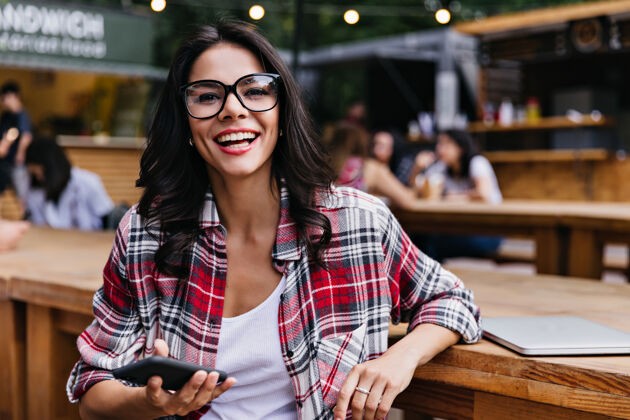 人时髦的白人女孩拿着智能手机微笑着在咖啡馆摆姿势美丽的女学生坐在户外餐厅与笔记本电脑自信笔记本电脑休闲