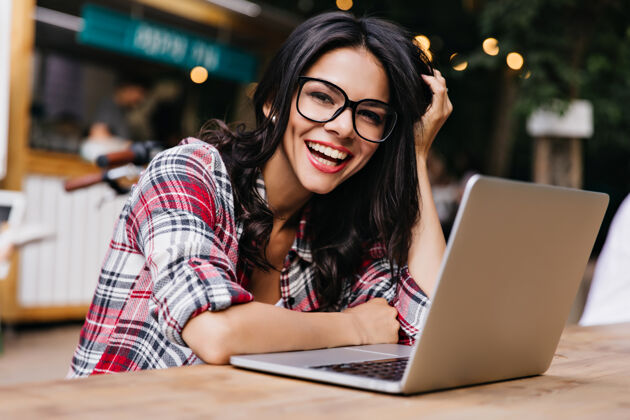 人迷人的黑发女青年在电脑前微笑戴着黑眼镜的情绪化女学生的户外照片教育女性自由职业者