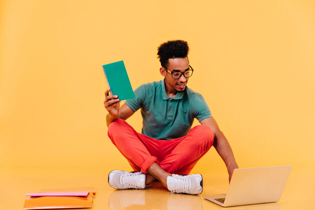 表情对眼镜感兴趣的非洲学生坐在地上拿着课本穿着鲜艳衣服的男性自由职业者使用笔记本电脑黄色模型国际