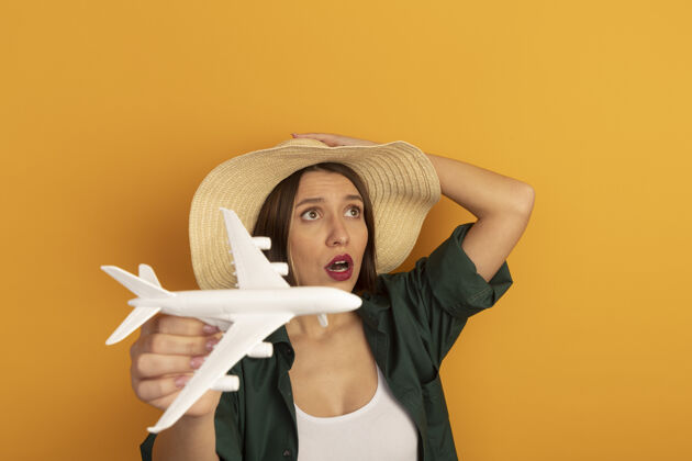 时尚带着沙滩帽的焦急漂亮的白种女人拿着模型飞机看着橙色的一面模特成年人黑发