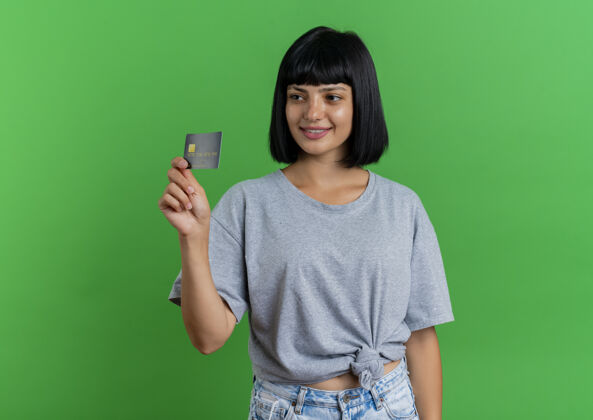 持有笑容可掬的年轻黑发白种女人拿着信用卡看着隔离在绿色背景和复印空间的一边信用壁板微笑