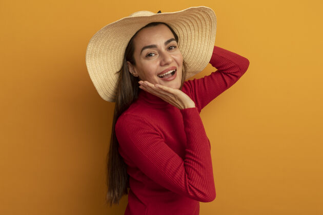 黑发快乐美丽的高加索女人 戴着沙滩帽 手放在脸上 看着橙色的相机快乐帽子时尚