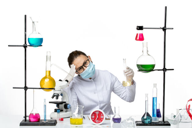 前面前视图：穿着白色医疗服的女化学家 白色办公桌上戴着面罩 化学家实验室病毒冠状病毒飞溅药物视图保持