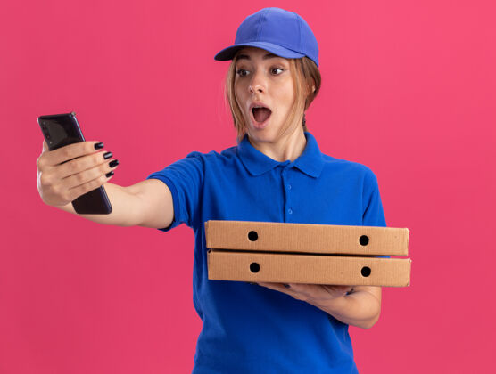 惊喜穿着制服的年轻漂亮的送货女孩拿着比萨饼盒 看着粉红色的手机姿势电话制服