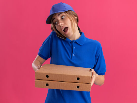 姿势给人印象深刻的年轻漂亮的送货员穿着制服拿着比萨饼盒 在粉红色的地板上讲电话披萨电话送货