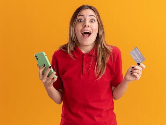 漂亮兴奋的年轻漂亮的白人女孩拿着信用卡和电话在橙色姿势信用成人