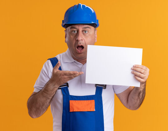 制服一个穿着制服的白人建筑工人拿着一张橙色的白纸 指着那张纸 大吃一惊建设者人成人