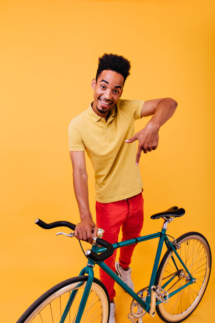 时尚一个穿着红裤子的笑脸黑人在玩的全长镜头一个骑自行车的活跃的非洲男人的室内肖像表情现代自行车