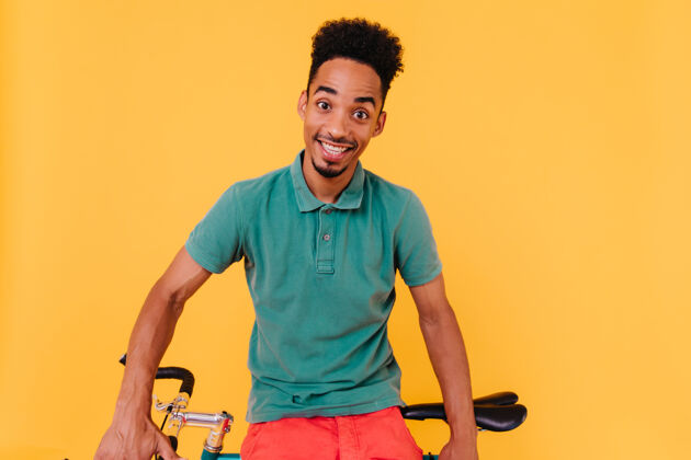 非洲一个穿着绿色t恤的骑车人带着惊讶的微笑摆出姿势坐在自行车旁边的一个惊讶的黑发男人的肖像黑色放松家伙