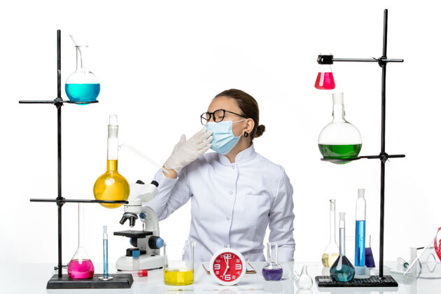 医学前视图：穿着医疗服的女化学家戴着面罩坐在白色背景上打呵欠病毒实验室化学冠状病毒飞溅前面坐科学
