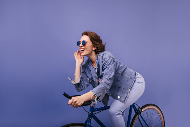 表情活跃的黑发女孩骑在自行车上室内照片欢快的女士在牛仔夹克坐在自行车上成人骑手牛仔裤
