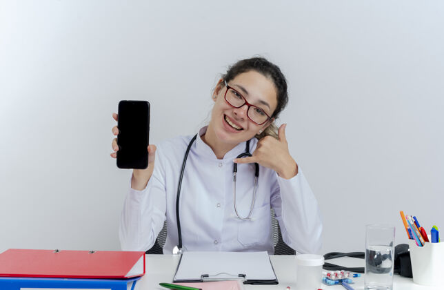 听诊器快乐的年轻女医生穿着医用长袍 戴着听诊器 戴着眼镜 坐在办公桌旁 拿着医疗工具 展示着手机 看起来在做通话手势欢乐手机医生