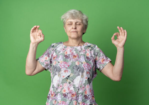 老人平静的老妇人假装冥想 双手孤立在绿墙上 打着ok手势手势老的冥想