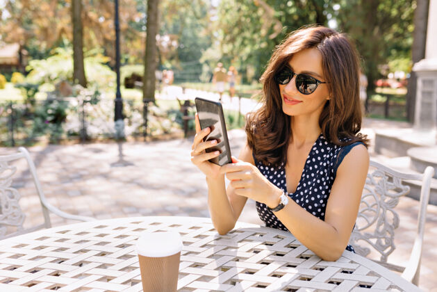 人一位留着黑色短发 笑容迷人的时尚美女 正拿着手机坐在阳光下的夏日自助餐厅里喝酒女太阳