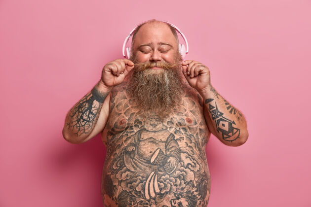 过量孤立镜头超重胡须男子卷曲胡须 闭上眼睛 听最喜欢的歌曲在耳机里 找到音乐站或有趣的播客 有纹身裸腹 模型对玫瑰色的墙壁大音乐心情