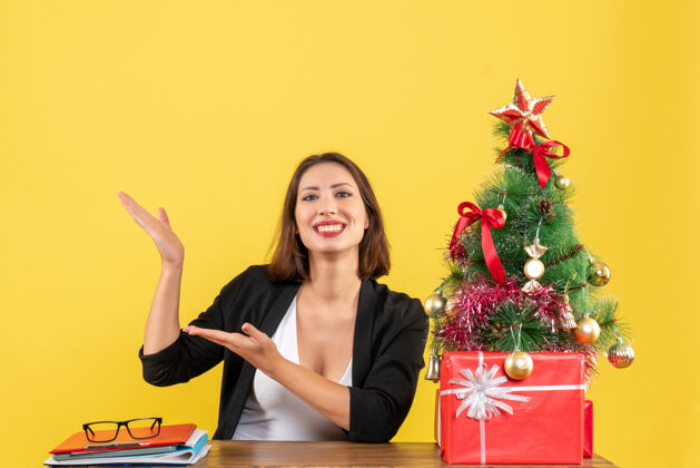 快乐一位年轻漂亮的女士在黄色办公室装饰的圣诞树旁的桌子旁自豪地展示着什么桌子封面女郎圣诞