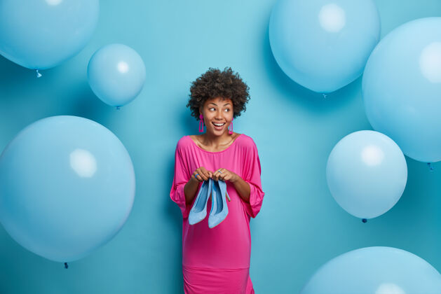 气球室内拍摄的是一个时髦的快乐女人 一头非洲头发 穿着粉红色的裙子 穿着高跟鞋 准备生日派对 试着选择穿什么 一边看 蓝色的气球在周围乐观节日年轻