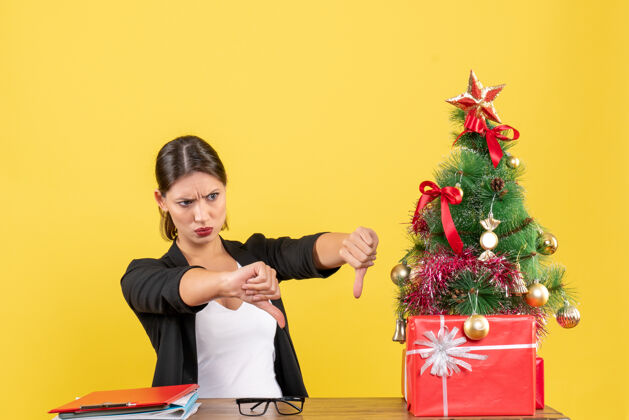 表演在黄色的办公室里 一个穿着西装的不开心的年轻女人在装饰好的圣诞树旁做着否定的手势消极生活手势