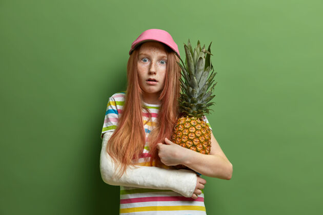 红发情绪恐惧的雀斑女孩拥抱菠萝 喜欢热带水果 戴帽子和条纹t恤 手臂骨折 隔离在绿色的墙上童年和生活方式的概念难以置信未成年多汁